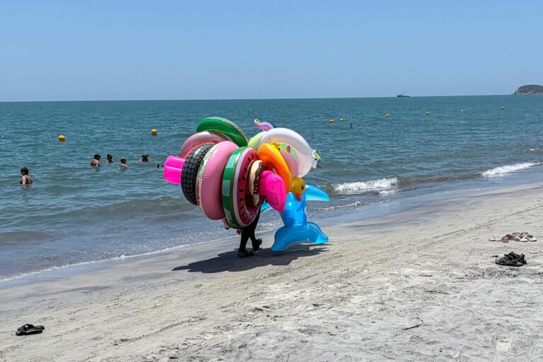 Playa de Santa Marta con turistas y vendedor de inflables, bajo un cielo soleado