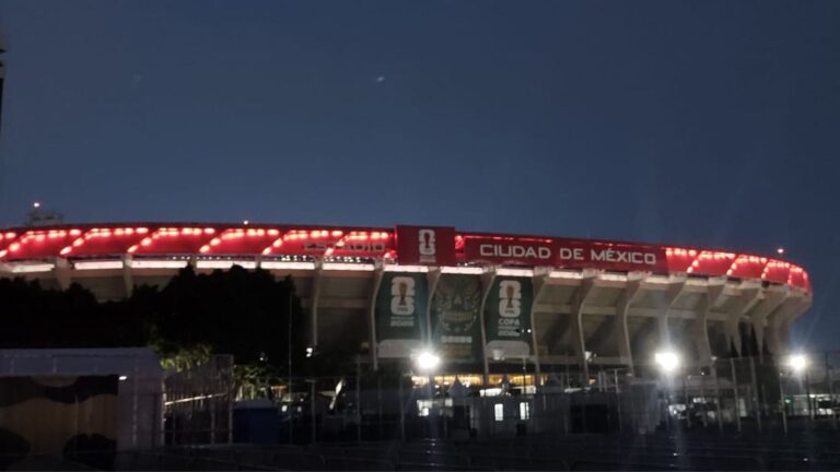 Vista del Estadio Ciudad de México iluminado con luces rojas al atardecer, con rótulos del Mundial 2026 en su fachada.